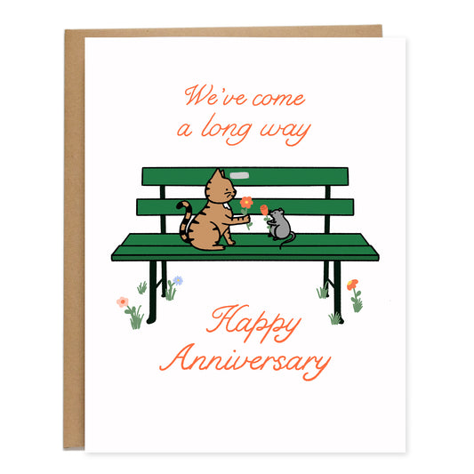 Card with a cat and rat holding flowers for each other sitting on a green park bench. The card reads, We've Come A Long Way Happy Anniversary in cursive lettering