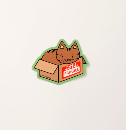 A green sticker with a drawing of a brown tabby cat inside of a cardboard box that has a red sticker on it that reads, handle with care fragile
