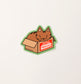 A green sticker with a drawing of a brown tabby cat inside of a cardboard box that has a red sticker on it that reads, handle with care fragile