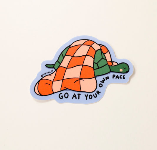 A drawing of a turtle sleeping with a fluffy, red and pink checkered blanket on top with a cornflower blue background. text on bottom reads, go at your own pace
