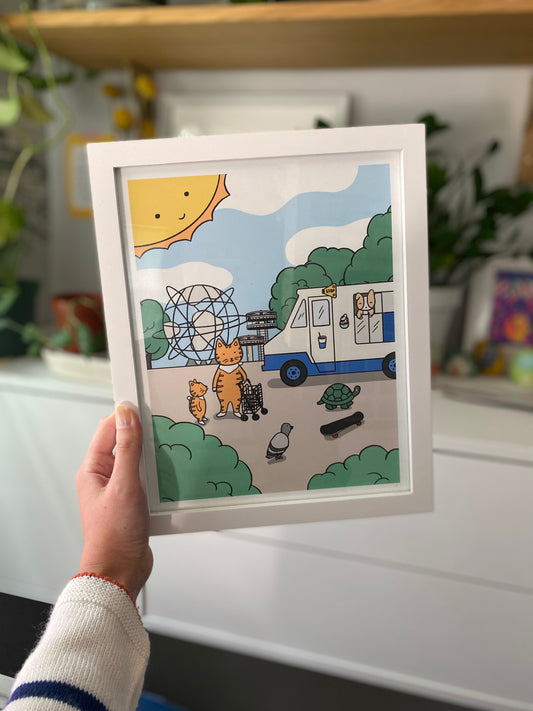 A hand holding up this art print, framed in front of blurry background of plants and shelves
