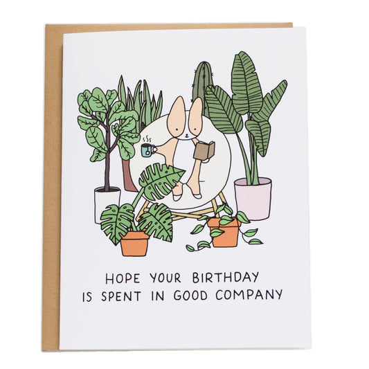 corgi surrounded by plants (monstera, fiddle leaf fig, bird of paradise) reading a book and drinking tea, card reads hope your birthday is spent in good company