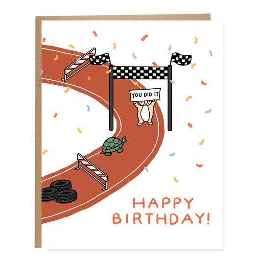 A card with a drawing of a red race track with hurdles and tires. A turtle is almost at the finish line with a corgi standing holding a sign that reads, you did it. Confetti is surrounding the race track. The card reads, happy birthday