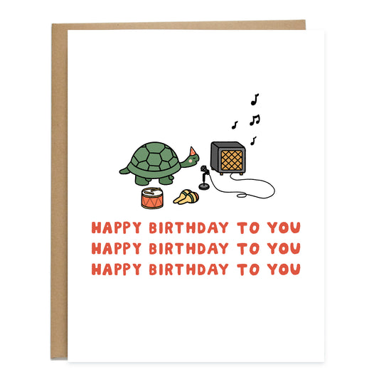 A birthday card with a drawing of a turtle singing in the middle into a tiny microphe that's connected to an amp. There's a tiny drum and maracas laying on the ground. The card reads, happy birthday to you, happy birthday to you, happy birthday to you, in bold, red text