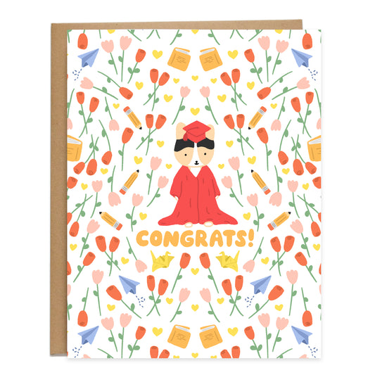 A tricolor corgi in the center wearing a red gown that's dragging on the floor and a red cap, surrounded by a pattern of red roses, pink tulips, yellow hearts, pencils, books, yellow paper cranes, and paper planes. Card reads, congrats, in orange bubbly handwriting