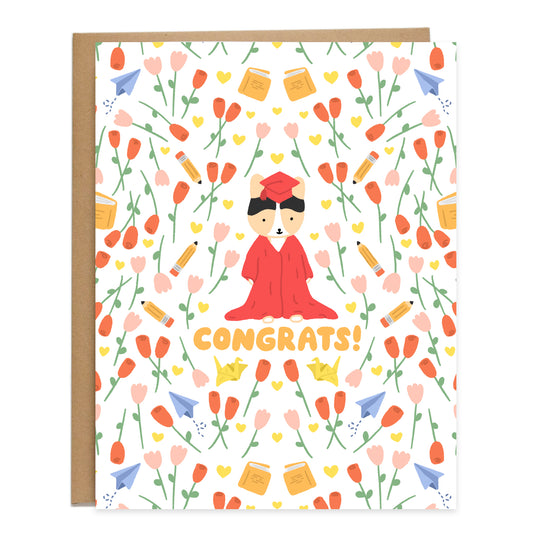 A tricolor corgi in the center wearing a red gown that's dragging on the floor and a red cap, surrounded by a pattern of red roses, pink tulips, yellow hearts, pencils, books, yellow paper cranes, and paper planes. Card reads, congrats, in orange bubbly handwriting