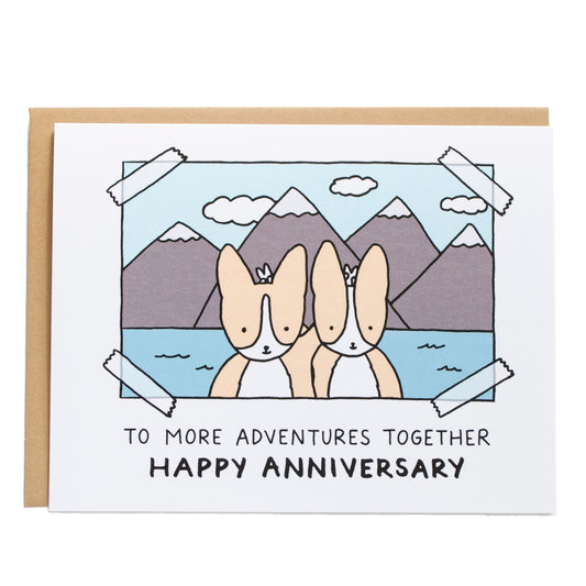 a taped photograph of two corgis putting bunny ears on each other in front of a scenic background of mountains, card reads, to more adventures together, happy anniversary