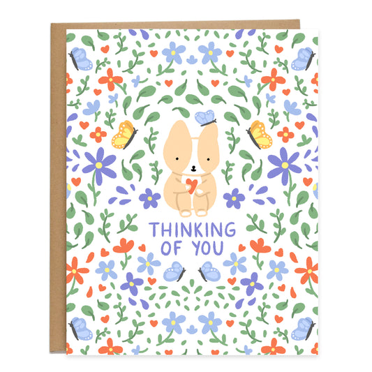 a drawing of a corgi sitting and holding its knees to their chest with a heart in their hand. a blue butterfly landed on it's ear. they are surrounded by a pattern of purple and blue flowers, leaves, yellow and blue butterflies