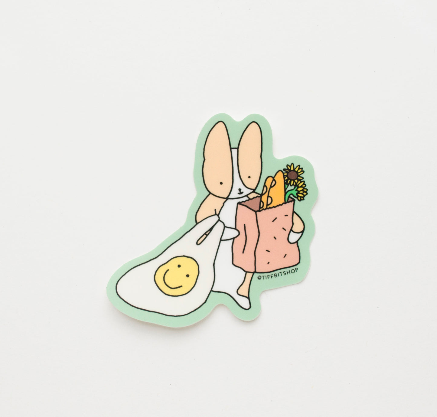 sticker with a green background of a corgi holding a reusable bag with a yellow smiley face and brown paper bag with baguette and sunflowers peeking out the bag