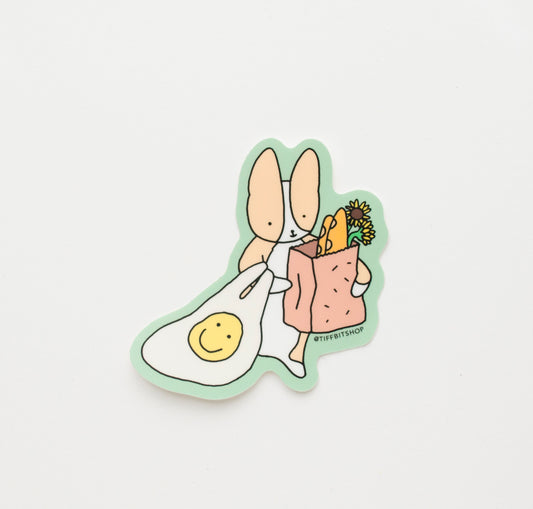 sticker with a green background of a corgi holding a reusable bag with a yellow smiley face and brown paper bag with baguette and sunflowers peeking out the bag