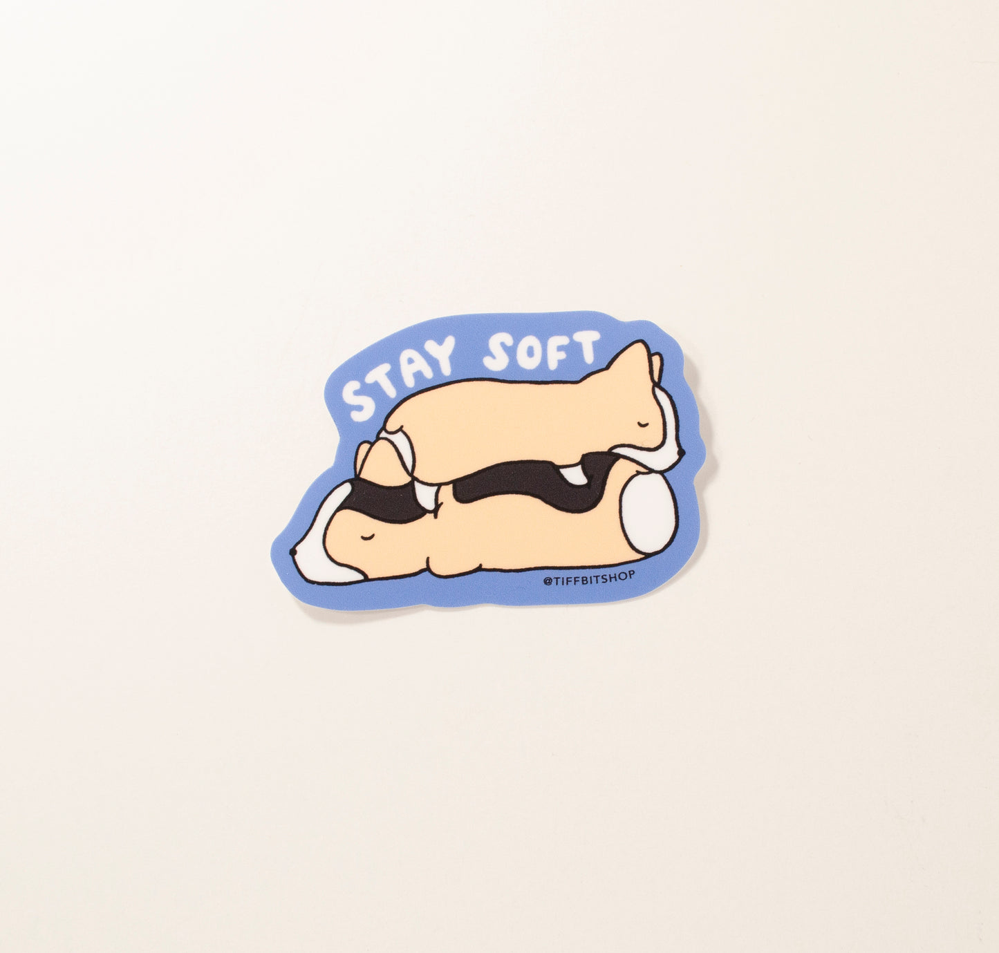 On a bright blue background, a drawing of two corgis stacked on top of eeach other sleeping. On top it reads, in white bubbly writing, stay soft