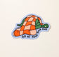 A drawing of a turtle sleeping with a fluffy, red and pink checkered blanket on top with a cornflower blue background. text on bottom reads, go at your own pace