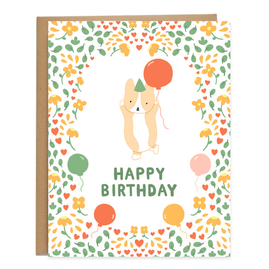 at the center of card is a corgi in the action of jumping with joy with its hands up, holding a balloon. surrounding the corgi is a pattern of flowers blooming, leaves, and balloons