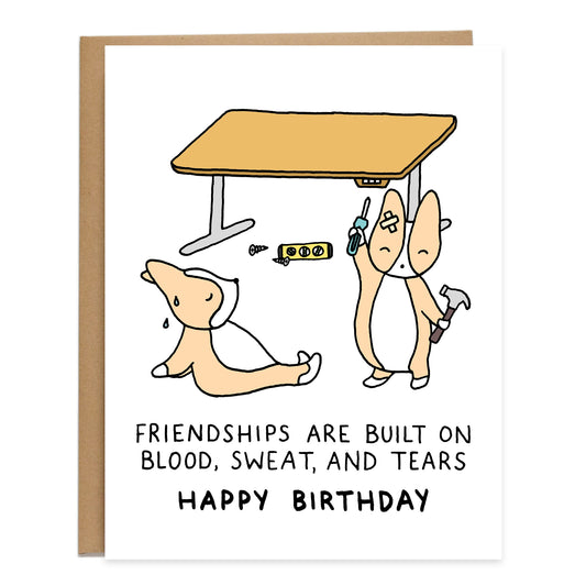 two corgis finished building a table, one corgi exhausted and the other standing holding up a screwdriver and hammer in their hands, text on card reads 'friendships are built on blood, sweat, and tears, happy birthday'