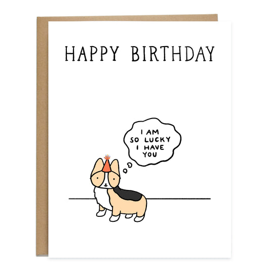 A birthday card with a corgi standing off to the side with a birthday hat on, a thought bubble reads, I am so lucky I have you. The card on top reads, happy birthday, in big letters