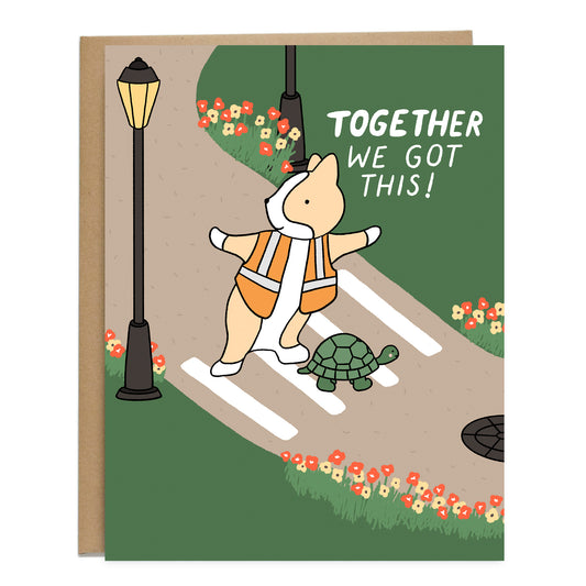 A card of a drawing of a turtle crossing a colorful street filled with flowers and grass and a corgi with its arms out wide, wearing a reflective jacket blocking any cars from coming. The card reads, together we got this!