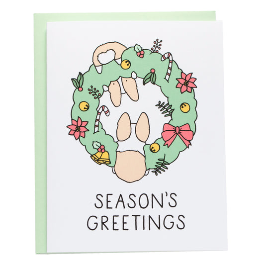 two corgis hanging from a green wreath covered with bells, bows, candy canes, and boughs of holly. card reads, season's greetings