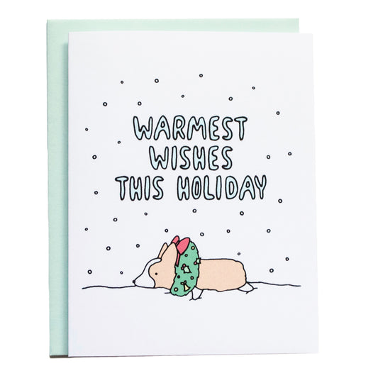 a corgi lying on snow with its head inside of a christmas wreath, card reads, warmest wishes this holiday