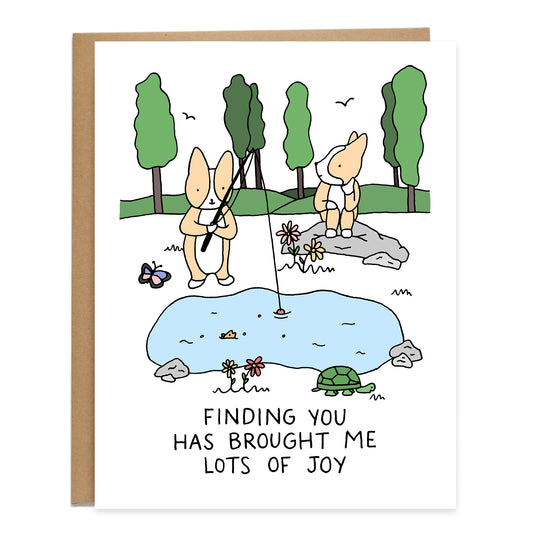 a scenic view of a corgi fishing and another corgi looking on sitting on a rock, trees in the background, flowers and small critters around them, text on card reads: finding you has brought me lots of joy