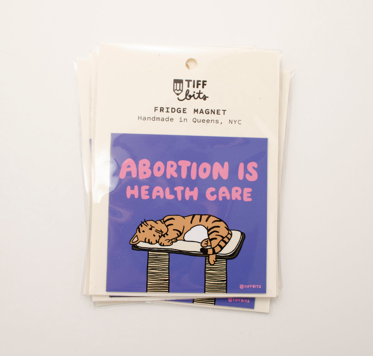 A magnet packaged in a clear bag featuring a drawing of a brown tabby cat laying on top of a cat tree, above it in pink letters reads, abortion is health care, against a purple background
