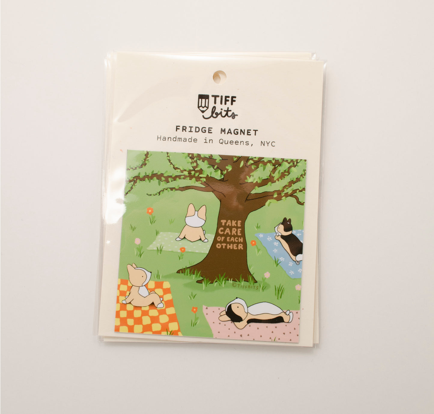 Magnet packaged in a clear bag featuring a drawing of four corgis laying on a grassy field, under a big tree, and each on a patterned blanket. On the tree reads, take care of each other