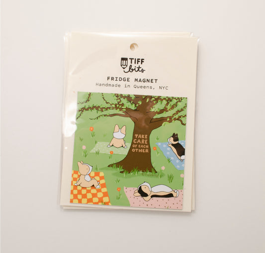 Magnet packaged in a clear bag featuring a drawing of four corgis laying on a grassy field, under a big tree, and each on a patterned blanket. On the tree reads, take care of each other