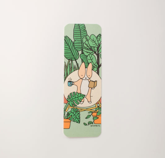 Bookmark design with rounded corners of a corgi sitting on a round chair reading a book in one hand and holding a mug of hot tea in the other, a group of houseplants surrounding it