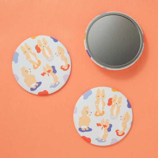 a photo of round pocket mirrors on a peach colored background. the design has five corgis standing on colorful blobs on a light blue background. one is posing with their hand on their hip, another is holding its head in their hands, one is standing with their hands behind their back, another is shaking its butt, and the last one has heart eyes popping out